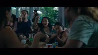 A group of Filipino women gather around a low, outdoor table. They are laughing, pointing and hitting the table. One woman stands up and smacks herself on the rear, and the laughter escalates.