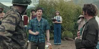 Hoa Xuande, wearing a blue plaid button-down shirt and holding a script, gestures to Robert Downey Jr. as he speaks. They are surrounded by members of the film crew on a jungle set.