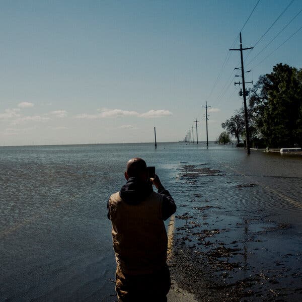 A man takes a photo as he stands at an intersection and looks out on a road covered in water and a landscape that looks like a sea.