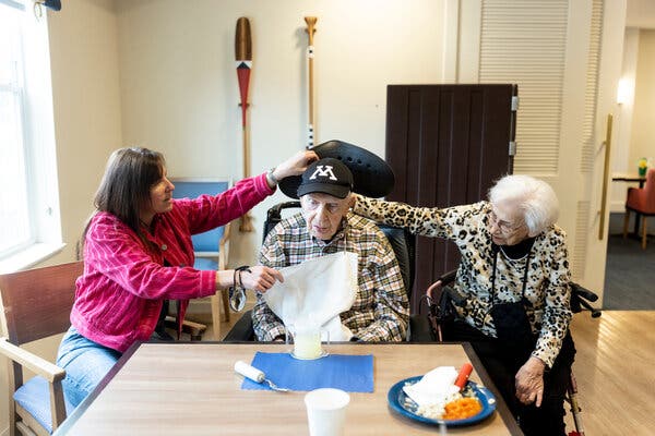 Anne Palm helps delicately remove a bib from the chest of her father, Donald Reiners, who sits at the head of a dining table in a wheelchair. He wears a plaid shirt and baseball cap. His wife, Florence, sits on his other side in her walker which is fitted with a seat, and she rests her hand on her husband’s back. A plate of half-eaten food rests on the table in front of them.