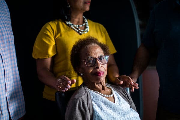 A portrait of Jewell Thomas, who sits in a chair with a white v-neck blouse and a gray sweater draped over her shoulders and white pearls, looking at the camera while her daughter, Angela Jemmott, rests her hands on the chair behind her, with her brothers standing on either side of her, faces out of frame.