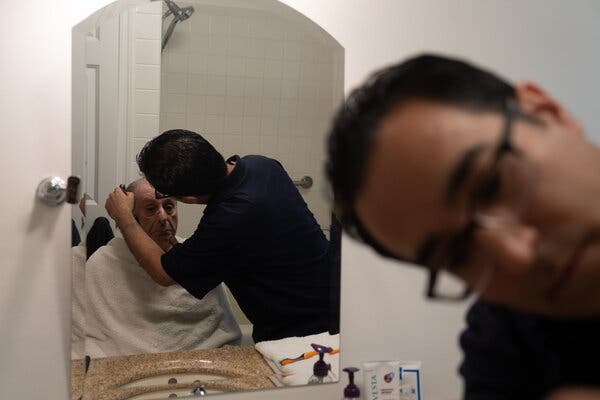 At right, a close-up of Robert Ingenito. Behind him in the reflection of a mirror, he can be seen shaving his father, Jerry, who is sitting with a white towel draped around him. 