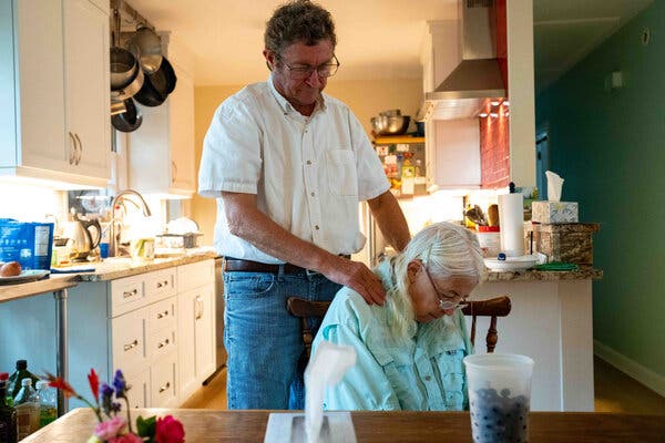 Robin Lee sitting at a table in a kitchen with her head down. Frank Lee is standing behind her and rubbing her shoulders. 