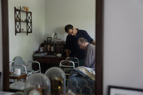 Through a door frame, Robert Ingenito can be seen standing and helping his father, Jerry Ingenito, getting out of bed. A walker sits in front of Jerry Ingenito. 