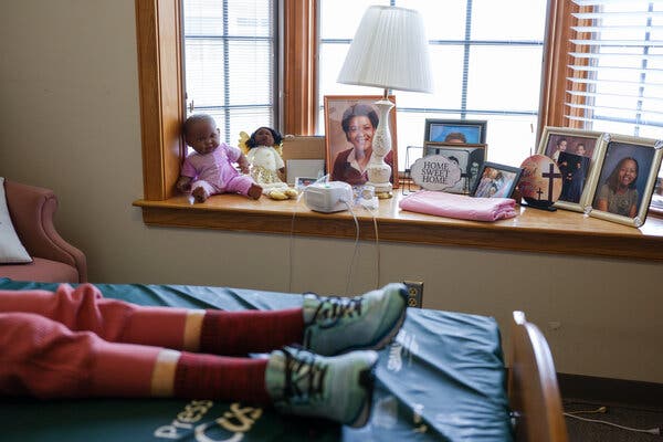 Two dolls dressed in pink and yellow clothes sit on a windowsill next to several framed photos. Betty Mae Glenn’s legs are stretched out on her nursing home bed. 