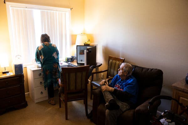 Jon Guckenberg looks on, seated in a large, brown plush chair in his room at an assisted living facility while his legal guardian, Nancy Pilger, her back to the camera, unpacks groceries to put in a mini-fridge resting on a table by a window.