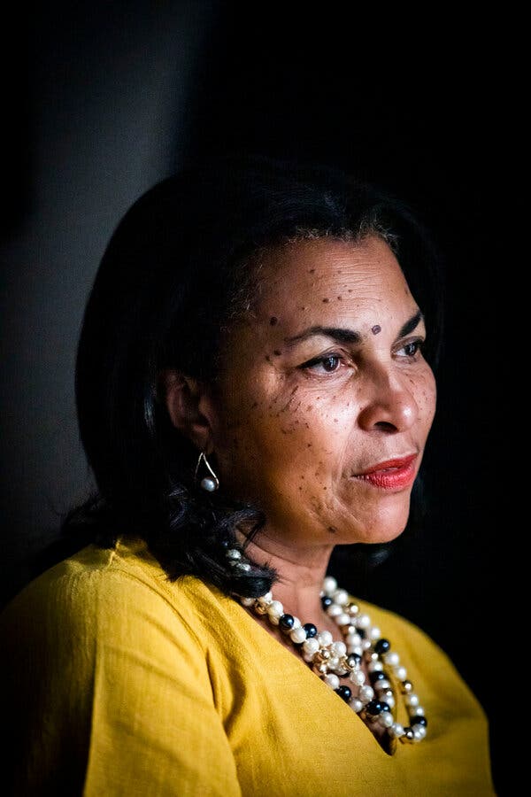 A portrait of Ms. Jemmott, who stands against a dark black background and wears pearls in her earrings and necklace and a yellow blouse as she looks off in the distance.