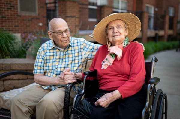 A portrait of Marlene Nathanson, who wears a bright red-pink shirt and a sun hat and sits in a wheelchair, next to her husband, Iric, who holds her hand and wears a blue and yellow checkered shirt.