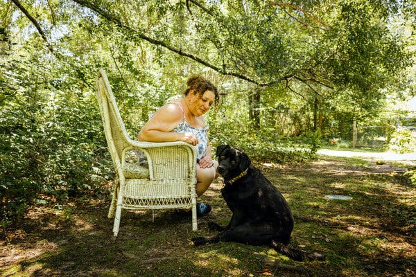 A woman in a floral dress sits under a leafy tree in a wicker chair beside a black dog that is sitting on the ground.