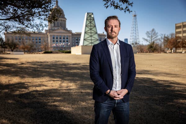 Ryan Walters, wearing a navy blazer over a white shirt and jeans, stands with his hands clasped in front of the Oklahoma Capitol.