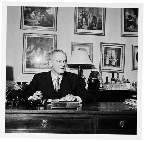 A black and white photo of Otto Lucas sitting behind a desk with framed pictures on the wall above him.