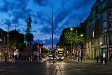 A city street at night. The sky is filled with dark blue clouds, and a person illuminated by car headlights crosses a road on a bicycle.