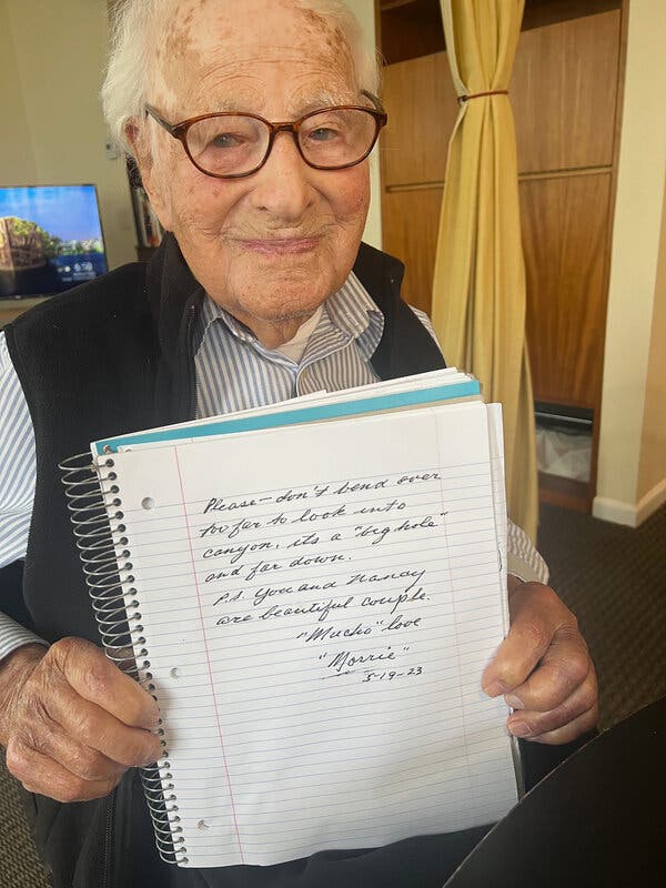 A photo of a more than 100-year-old man wearing a vest, striped shirt and glasses, holding up a spiral-bound notebook with a handwritten note on the page.