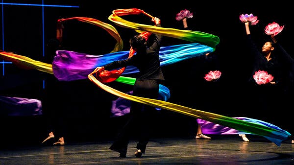 A dancer in a spotlight twirls while holding rainbow-colored ribbons against a darkened backdrop.