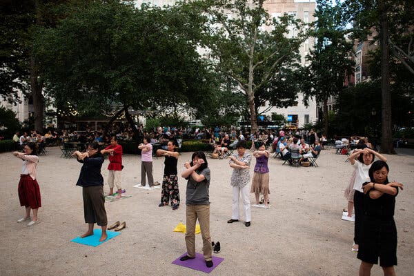 A group of people stand spaced apart from one another while holding their arms together in front of their bodies outside in a park. 