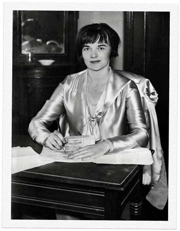 A black and white portrait of Ursula Parrott sitting at a table with a pencil in her right hand and looking up at the camera with a slight smile.