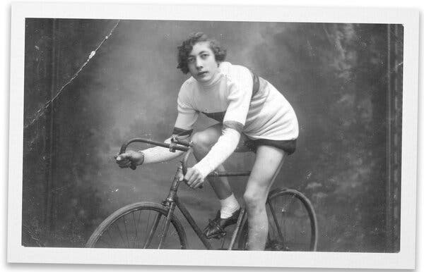 A black and white studio portrait of de Bruyn in a long-sleeved white riding jersey and dark shorts sitting on his bicycle and leaning forward while gripping the handlebars. His curly hair, parted on the side, covers his ears.