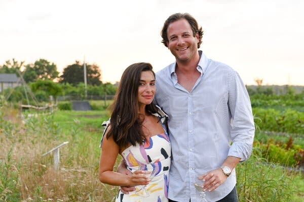 Candice Miller, in a white summer dress, poses for a portrait with her husband, Brandon Miller, in a light blue shirt. Both are holding wine glasses.