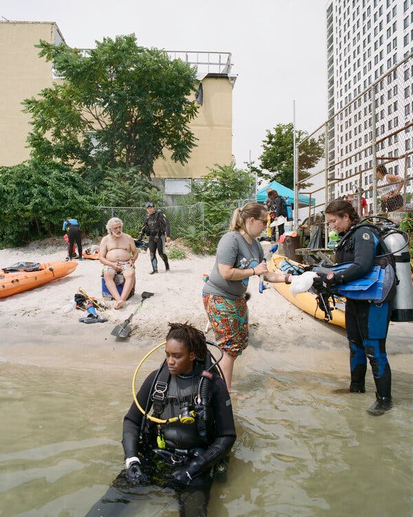 A scuba diver sits in shallow water on a beach filled with other people, some of whom are wearing diving gear.