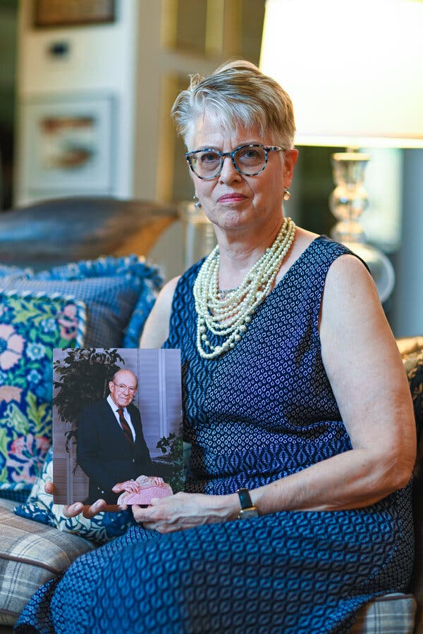 Beverly Brandt wears necklaces and a blue patterned sleeveless dress while sitting on a couch and holding a photo of her stepfather.