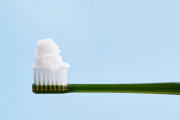 A photo illustration of a green toothbrush with coconut oil piled on top of the bristles. The background is a light blue. 