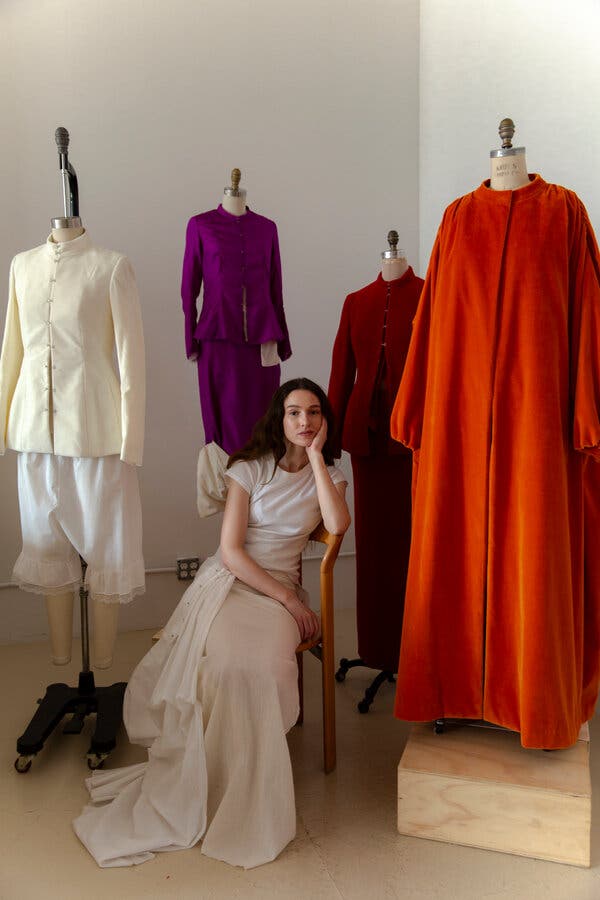 Colleen Allen sits on a chair with her left elbow on the arm, and her cheek in the palm of her hand. Four outfits in cream, violet, red and orange are displayed on mannequins around her.