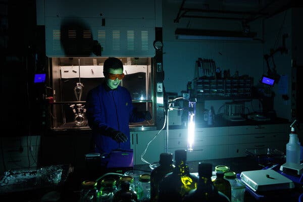 In a darkened lab, a man wearing red safety glasses and a blue lab coat looks at a source of intense light. 