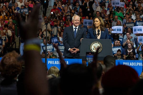 Tim Walz smiles broadly with his hands clasped before him, standing next to Kamala Harris. The two are on stage at a packed rally with people holding signs.