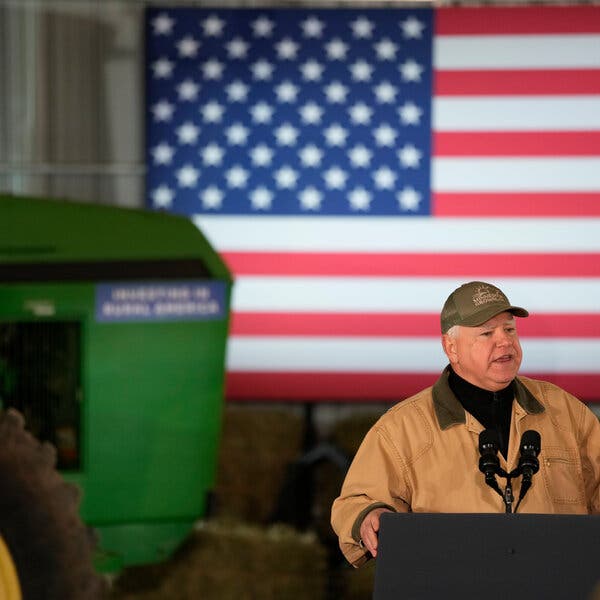 Tim Walz speaking at a lectern with a large American flag in the background.