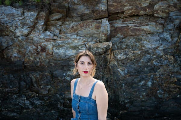 Daisy Alioto, wearing bright red lipstick and a denim tank dress, in front of a large rock.