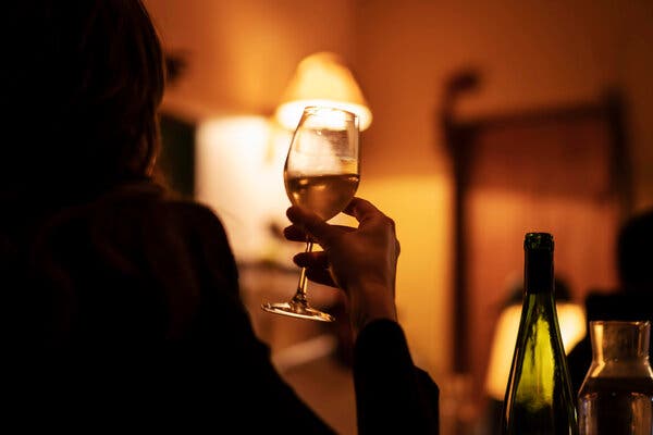 A view from over a person’s shoulder. The person is lifting up a full glass of wine with their right hand in a softly-lit wine bar.