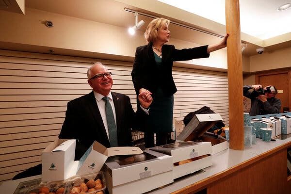 Ms. Walz, wearing a black pantsuit, stands up above a table covered in boxes of doughnuts, holding her husband’s hand.