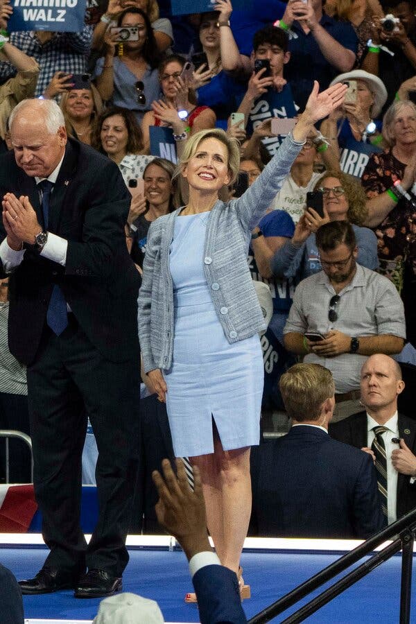 Gwen Walz, wearing a blue dress with a cardigan, waves from a stage alongside Tim Walz, who’s wearing a dark suit.