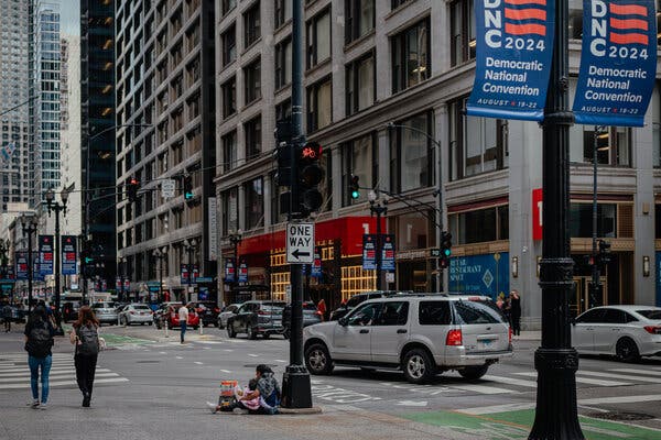 An adult sits on a busy street corner as people and cars pass by. 