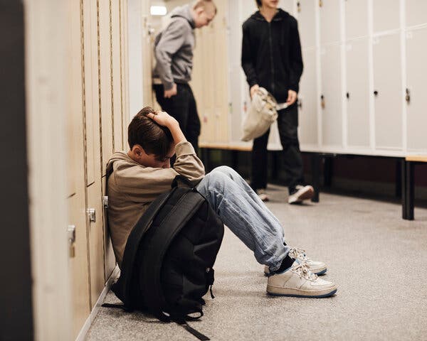 Boy sits behind his backpack leaning against lockers with his hand on his head while two other people walk down the hall.