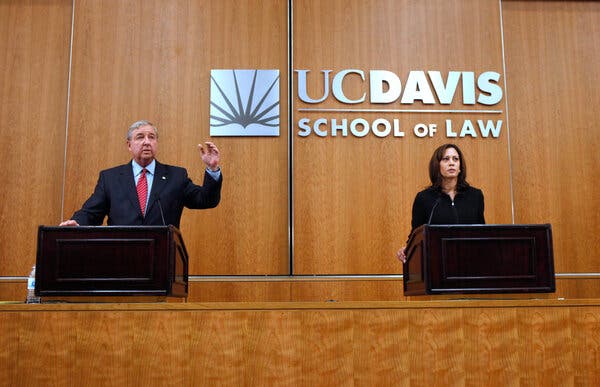 A man, gesturing, and a woman stand behind podiums and in front of a wood panel wall that reads, ‘UC Davis School of Law.’