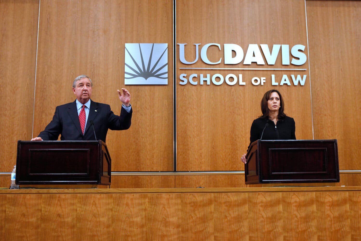 A man, gesturing, and a woman stand behind podiums and in front of a wood panel wall that reads, ‘UC Davis School of Law.’