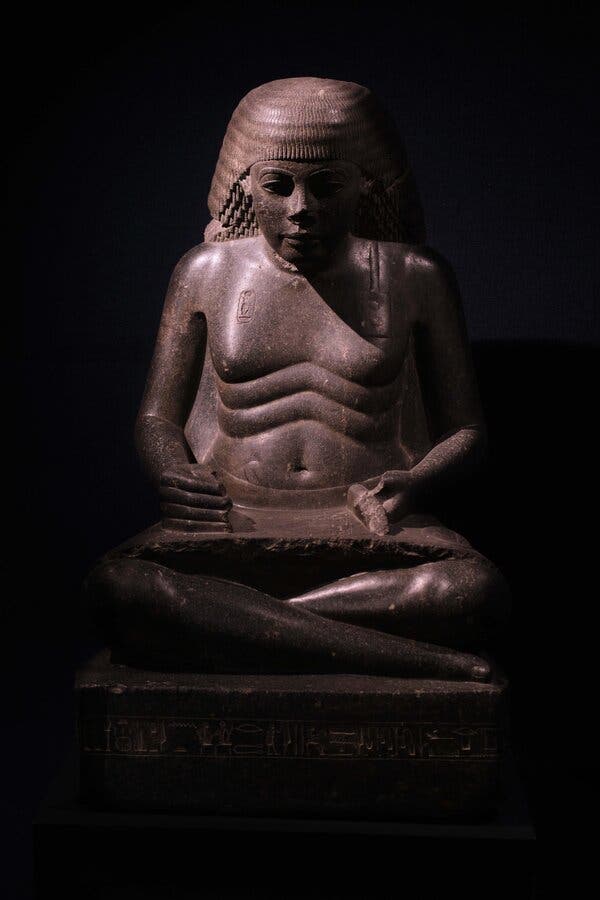 A statue of a scribe in a dark museum space, sitting cross-legged with a tablet in his lap and his right hand in a stylus-grasping position.