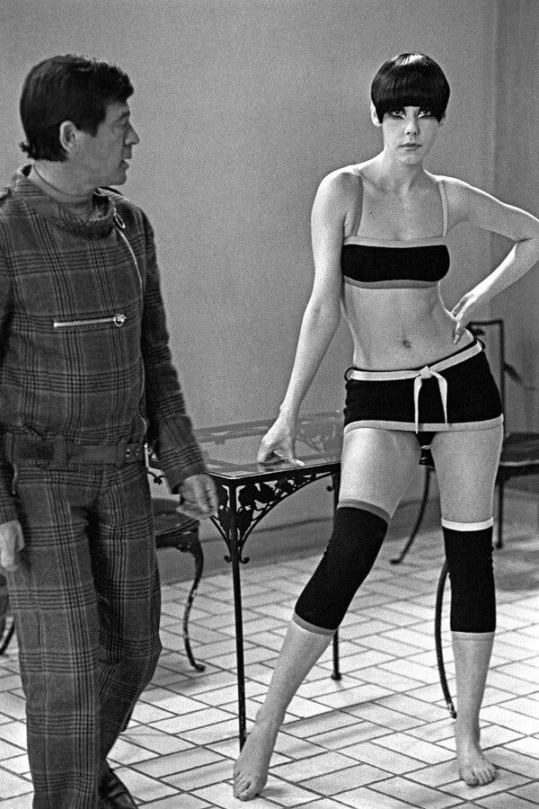 A black-and-white photo of Ms. Moffitt, wearing a bikini and knee bands, standing next to Rudi Gernreich, a man dressed in plaid and photographed in profile. She has her left hand on her hip and her right hand on a glass table. She wears her hair in bangs and is barefoot.