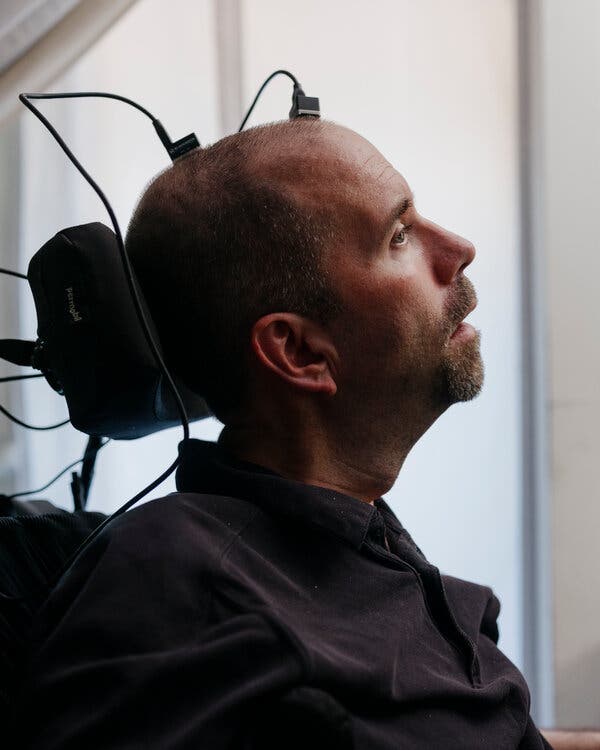 Casey Harrell, with his face facing right, sits in a wheelchair, with two portals attached to the top of his head and cords leading from each of them.