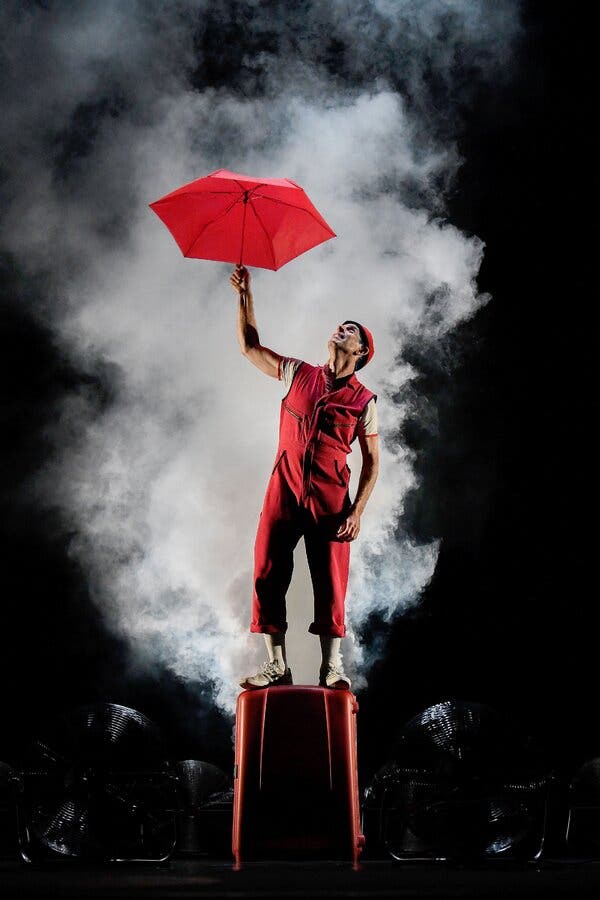 Seth Bloom, wearing a red jumpsuit over a T-shirt, looks up at the open red umbrella he is holding over his head. He is standing on a red platform on a dark stage. A cloud of white smoke billows behind him.