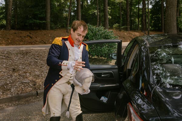 A man dressed as the Marquis de Lafayette, standing next to his car, puts his wig on a holding stand.