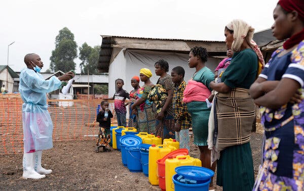 A health worker in boots and hazard wear stands in front of a row of women and children with buckets on the ground in front of them near a building.