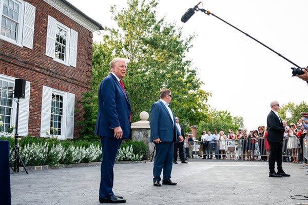 Former President Donald J. Trump, wearing. a blue suit and red tie, stands outside a brick building. A microphone is in front of him, and a group of people behind a barrier is to his side.