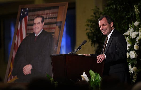 John Manning speaks at a podium next to a portrait of Justice Antonin Scalia.