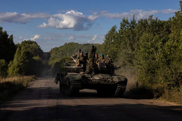 Ukrainian troops in military uniform sitting on top of tank as it drives down a dirt road flanked by trees.