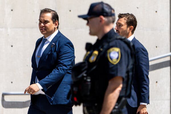 George Santos in a blue suit leaves Federal District Court in Central Islip, N.Y., earlier this week. A law enforcement officer is in the foreground and Mr. Santos is followed by another man in a suit.