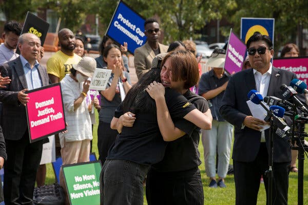 Two people dressed in dark clothing embrace in the foreground as other people gather around them, some holding signs with slogans like “Demand Justice, Demand Truth.”