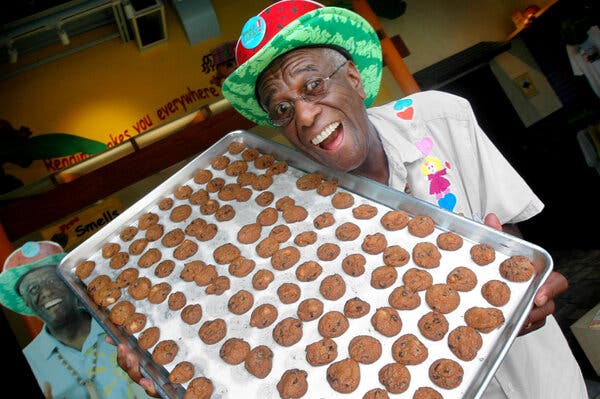 Smiling broadly as he looks at the camera, he wears a bright green and red patterned Panama hat while holding a large aluminum tray of baked cookies.