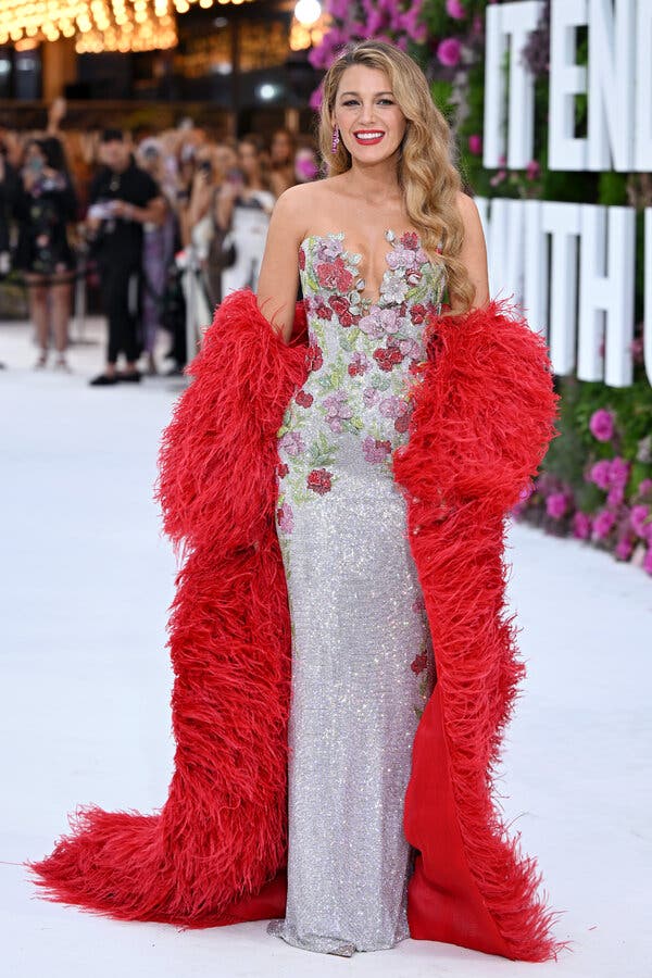 Black Lively poses on a white carpet in a long glittery dress adorned with fabric flowers and a red boa. Letters spelling out “It Ends With Us” amid real flowers are blurred in the background.